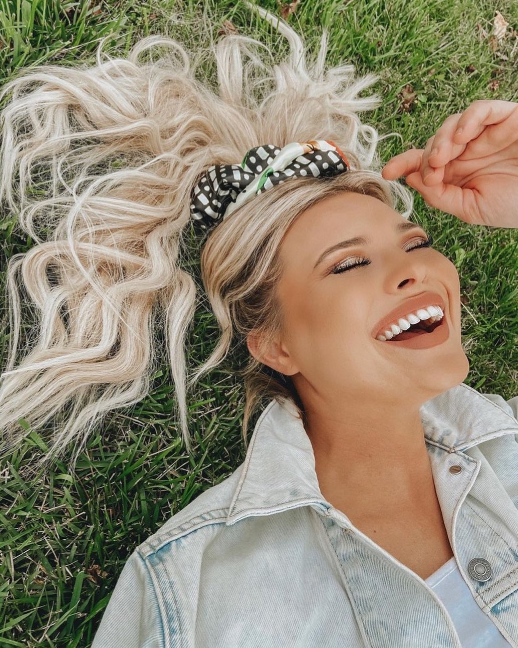 blonde Woman wearing giant scrunchie laughing laying in the grass. Wearing a print and denim jackt